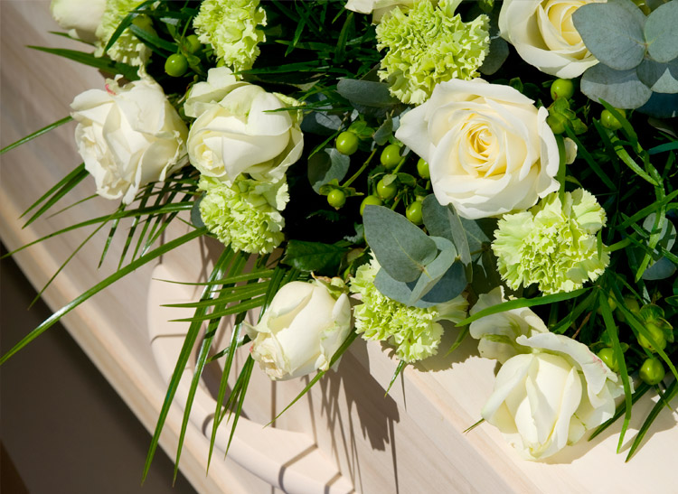 Bouquet of white roses and green carnations resting on an ornate, light-colored wooden casket. The arrangement is accented with green berries, long foliage, and eucalyptus leaves.