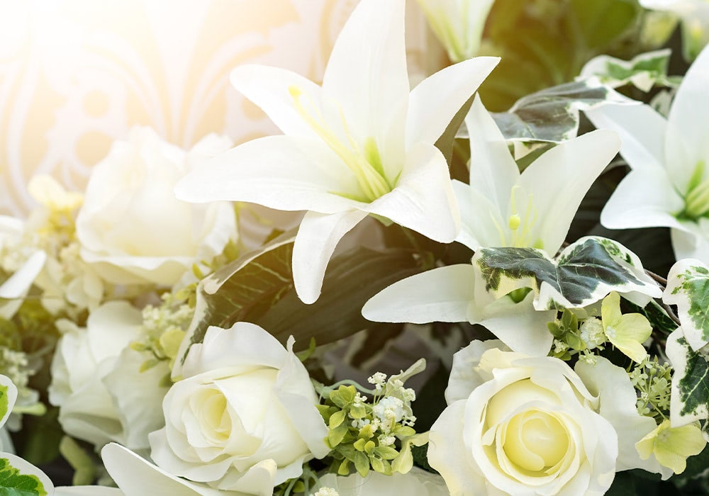 White lilies and roses in full bloom are arranged in a bouquet, set against a light backdrop surrounded by green leaves and smaller, delicate blooms.